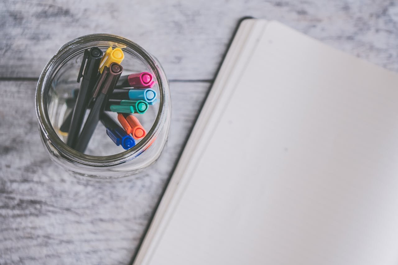 Coloured pens in a jar beside a blank notebook